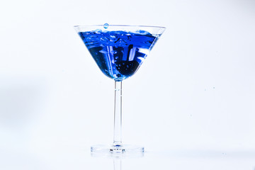 Cocktail with blue liquid in glass. Glass with blue water pouring with liquid with splashes and drops. Martini glass filling with alcohol with splashes on white background. Refreshing drink concept.