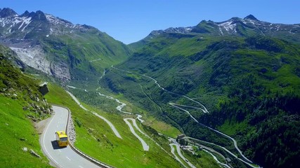 Swiss yellow bus driving on serpentine from right to left while camera moves slowly towards the pass with large mountains in the far.