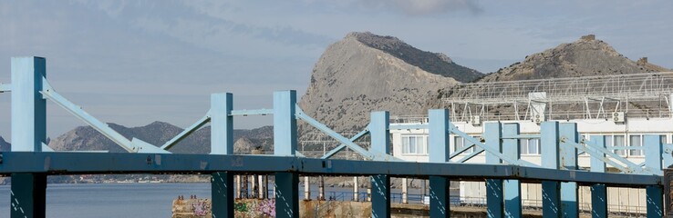 Scenic panoramic view of Sudak coastline behind large metal constructions on beach embankment towards Fortress and Hawk mountains and New World location, Crimea, Russia.