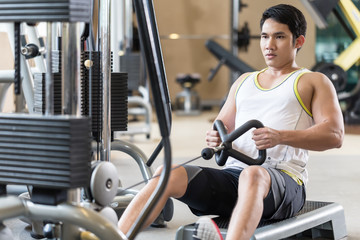 Portrait of a determined handsome young man looking forward while rowing at the cable machine during workout for back muscles in a modern fitness club