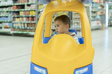 Cute sad little 2 year old baby boy child in the little toy-car trolley at supermarket, Dad or Parent pushing child shopping cart car with his son, Kid first experience concept