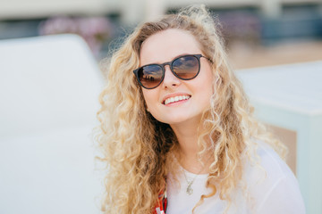 Photo of attractive young female has light curly hair, wears trendy shades, has toothy smile, shows...