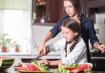 Cute little girl and her beautiful mom are cutting fruits, red watermelon and smiling while cooking in kitchen at home. Happy Family