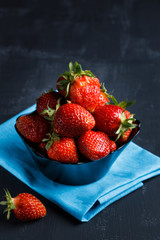 Ripe strawerry in a black plate on a blue napkin. Blurred dark background. The concept of a healthy diet.