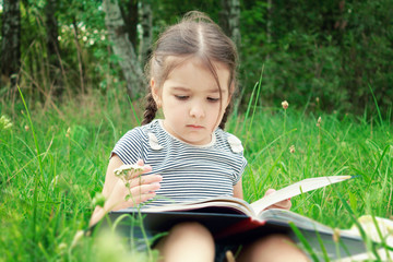 A girl in summer sits on the grass and reads a book outdoors