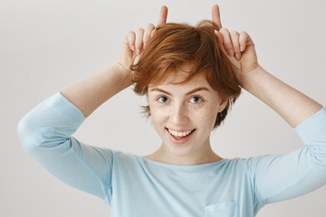 Every redhead have little devil inside. Close-up portrait of positive good-looking caucasian girl with ginger hair holding index fingers as if it is horns behind head, smiling broadly at camera