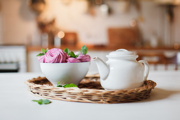 White teapot and pink dessert are serving on straw wicker tray on table. Tea time, cozy atmosphere. Kitchen still life on background with warm light. Sweets are decarated with herbal mint leaves.