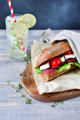 sandwich with salami, brie cheese, lettuce and cherry tomatoes on a wooden plate, a glass of lemonade with lime, ice and thyme. selective focus and copy space