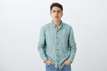 Indoor shot of childish displeased european guy in casual outfit, holding hands in pockets, frowning and showing tongue at camera, expressing dislike and disdain, standing over gray background