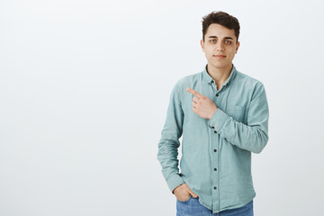 Portrait of calm confident good-looking boyfriend in shirt and jeans, holding hand in pocket and pointing at upper left corner with slight smirk, being self-assured and pleased over gray wall