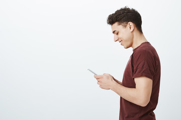 Profile shot of positive good-looking boyfriend in red t-shirt, holding smartphone and smiling at screen while typing message or searching infortmation in internet, standing happy over gray wall