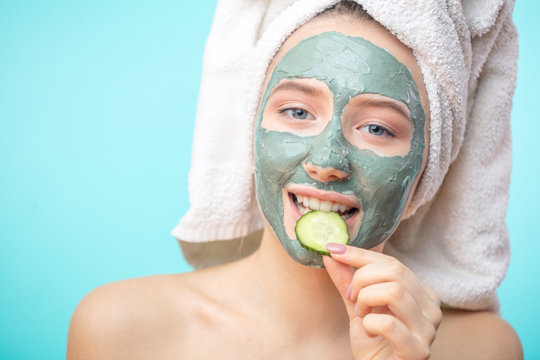 Funny young girl in positive cheerful mood with her haie wraped in towel holding slice cucumber for eyes treatment isolated over blue background.