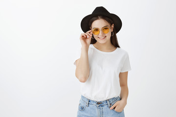 Girl getting ready for awesome party, dressing up in stylish hat and sunglasses, touching rim and smiling broadly, holding hand in pocket, feeling confident and amused, spending time with friends