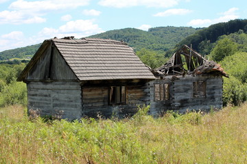Old wooden house ruins with partially broken and fallen roof, missing windows and dilapidated wooden boards surrounded with tall uncut grass with forest vegetation and cloudy blue sky in background