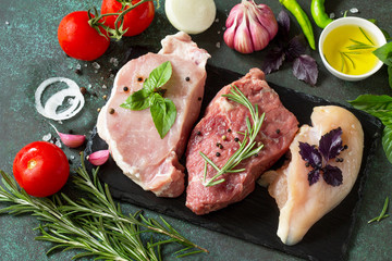 Fresh raw meat. Different types of raw pork meat, chicken fillet and beef with vegetables and herbs on dark wooden background.