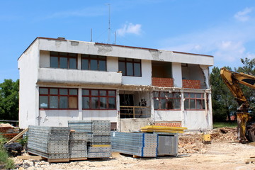 Small white building reconstruction with destroyed windows, doors, balcony and construction material and rubble in front and cloudy blue sky in background