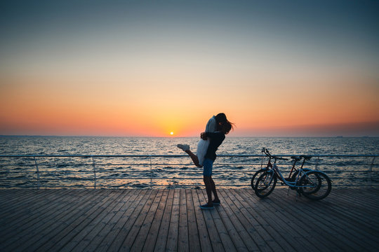 Man holding his girlfriend, couple of young lovers kissing at the beach at sunrise sky at wooden deck summer time