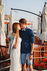 Cute young beautiful couple kissing near to the small summer cafe at port, happy smiling outdoor portrait