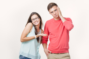 Нoung couple standing together over white background. Smiling woman points a hand at a man with a surprised and unhappy expression on his face. Discord in the relationship