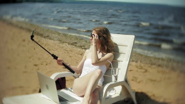 A young girl in a white bikini lies and sunbathes on a deckchair on a sea sandy beach and photographs on a smartphone SELFI