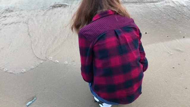 young woman dressed in jeans and a plaid shirt is enjoying a stay on the coast, overlooking the sea and the waves, playing with sand