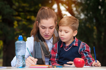 Positive cheerful young woman, mother is engaged with her little son developing activities sitting at a table in the courtyard of a private house against the background of blurry trees