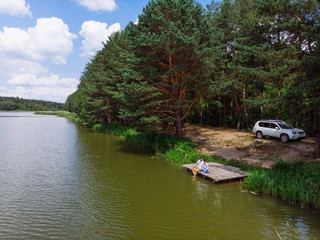 Fototapeta na wymiar aerial view couple sitting at wooden doc looking at lake. suv on background