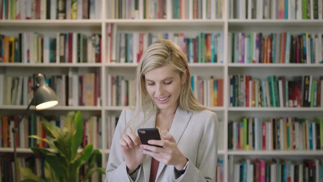 portrait of stylish blonde business woman smiling enjoying texting browsing online using smartphone social media app wearing suit in library office