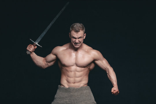 Bodybuilder man posing with a sword isolated on black background. Serious shirtless man demonstrating his mascular body