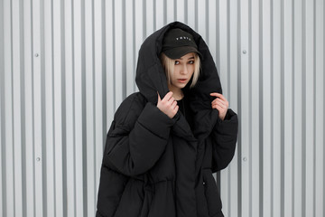 Naklejka premium Fashionable beautiful model woman in black fashionable winter jacket and stylish cap posing near metal wall