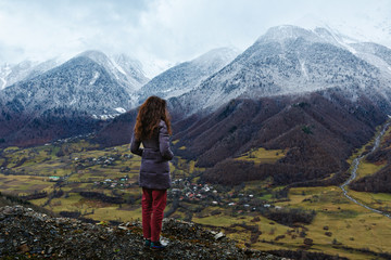 Naklejka premium Young woman stands over a cliff and looks at snow-capped mountains, village in lower mount. Winter is coming, first snowfall, change of seasons. Georgia.