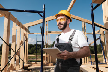 The man is a builder on the background of the roof of a frame house, in a yellow helmet and gray overalls. Uses a tablet