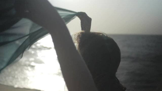 joyful girl is playing with flying silk scarf in sunny windy weather in a sea beach, against the sun