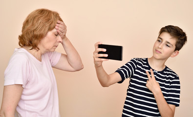Son taking selfie while his dissatisfied mother is slapping her head with hand. Family and modern technology addiction concept.