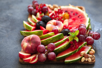 Fruit salad with watermelon, plum, peach, red currant, grape and nuts on wooden tray over dark slate background