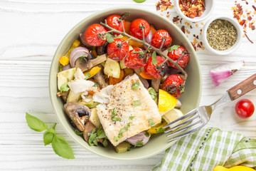 Baked vegetables with mushrooms and white fish fillets in bowl. Zucchini, cherry tomatoes on a branch, red onions, bell peppers and garlic. Healthy diet food