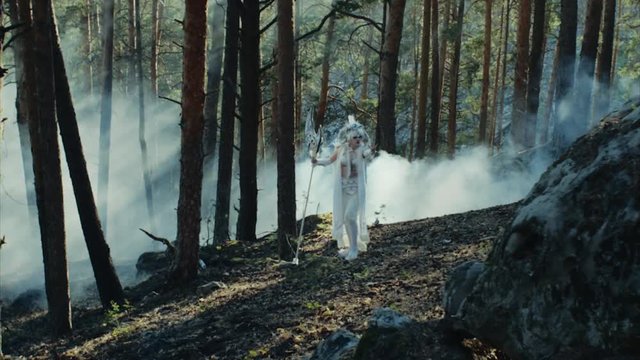 Satyr in white robe with ax axe and horns on the head appeared in the forest among trees. Wood spirit, smoke on the background. Empty wood.