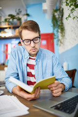 Young analyst or economist sitting by table in cafe with laptop in front and reading book