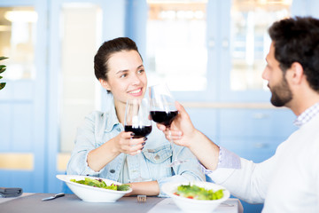 Young amorous couple clinking with red wine while celebrating family occasion in restaurant