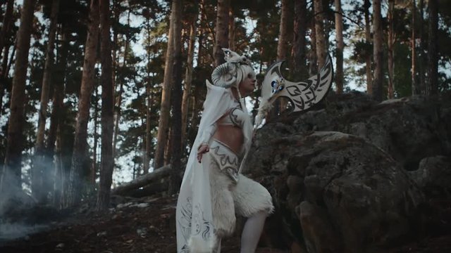 Model dressed like mythical faun is strolling in forest near spruces. She is carrying hatchet in hands, tilt up view