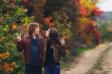 cheerful couple shows emotions. man and woman in leather jackets and jeans show each other tongues against background of autumn trees.