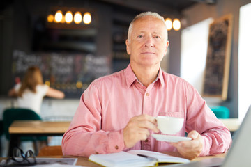 Serious senior man with cup of tea sitting by table in cafe while planning work at coffee-break