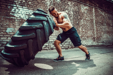 Muscular bearded tattooed fitness shirtless man moving large tire in street gym. Concept lifting,...