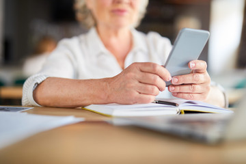 Senior businesswoman with smartphone looking through contacts or texting by table