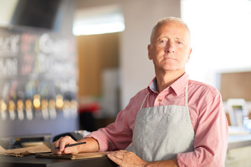 Aged man in apron looking at camera while waiting for new guests by bar counter in cafe or restaurant