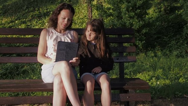 Family with a tablet on the bench. Mother with daughter with gadget in the park.