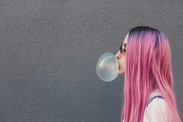 Stylish young hipster woman with long pink hair blowing a bubble with bubble gum.