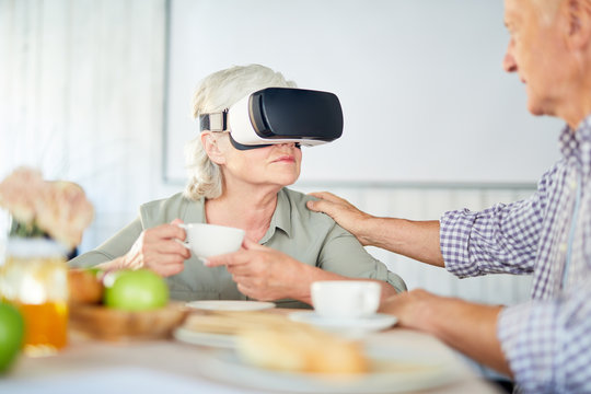 Senior husband comforting his wife wearing vr goggles during talk by cup of tea
