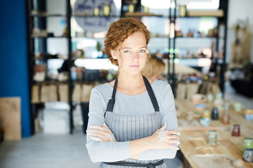 Young craftswoman with her hands in clay standing in workshop with colleague on background