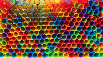 Hundreds of colorful plastic straws laying facing forwards opening towards viewer, laying on yellow surface. Many cities are now banning single use plastic straws.